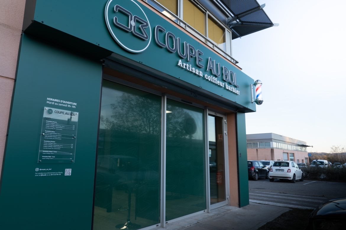 Façade verte du salon Coupe au Bol, avec l’enseigne « Artisan coiffeur barbier » et le poteau de barbier