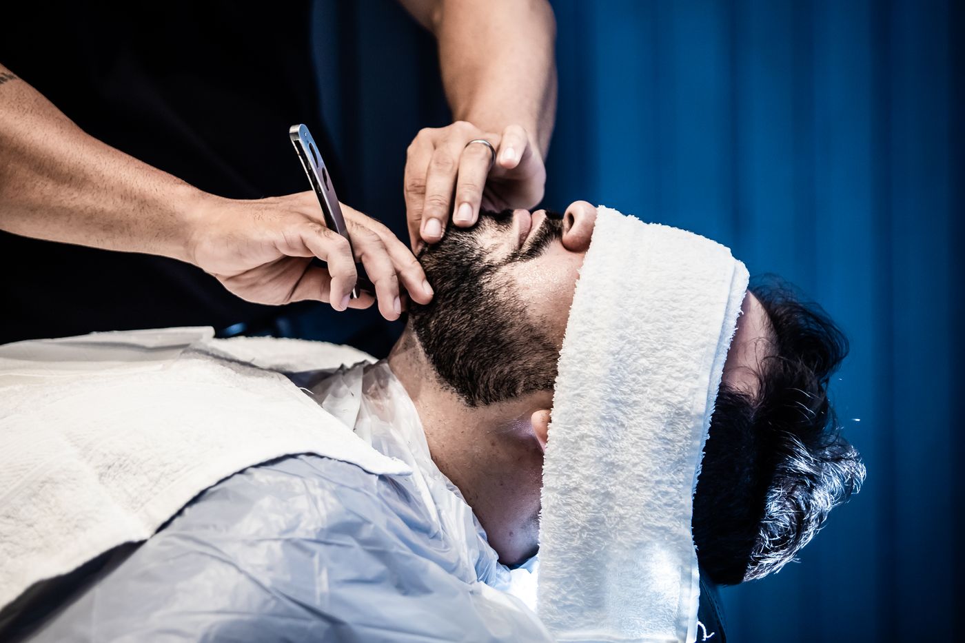 Un barbier taille la barbe d’un client allongé, une serviette chaude posée sur le front